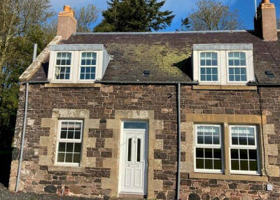 A two-story residential house with a gray stone facade, white window frames, and a black roof, set against a blue sky with scattered clouds.